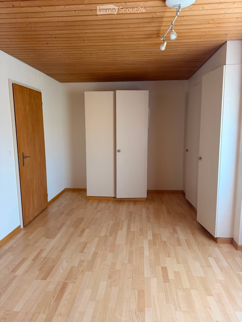 Empty hallway with light wooden flooring, white walls, and a wooden door on the left side. The ceiling is also made of light wood with a modern light fixture.