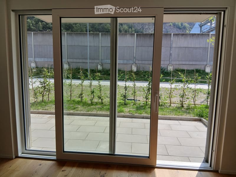 Sliding glass door opens to a tiled balcony area with small plants and a concrete wall in the background.
