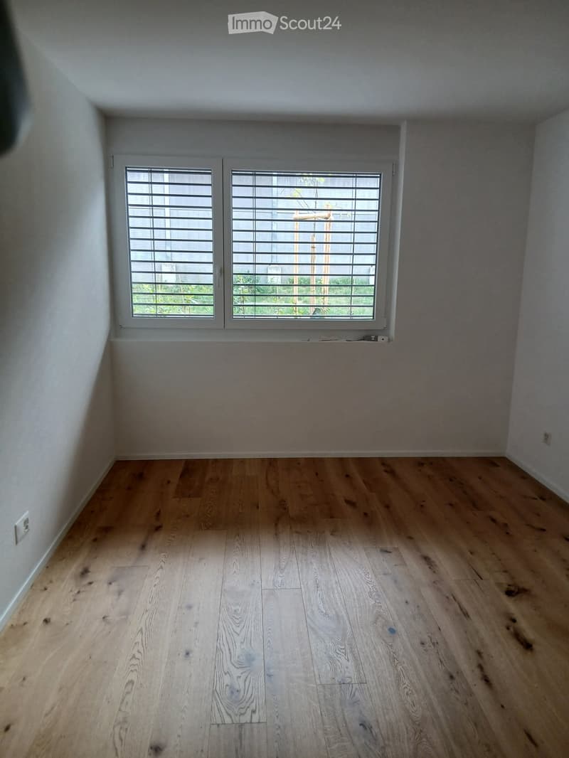 Empty room with light wooden flooring and white walls, featuring a large window with blinds offering a view of greenery outside.