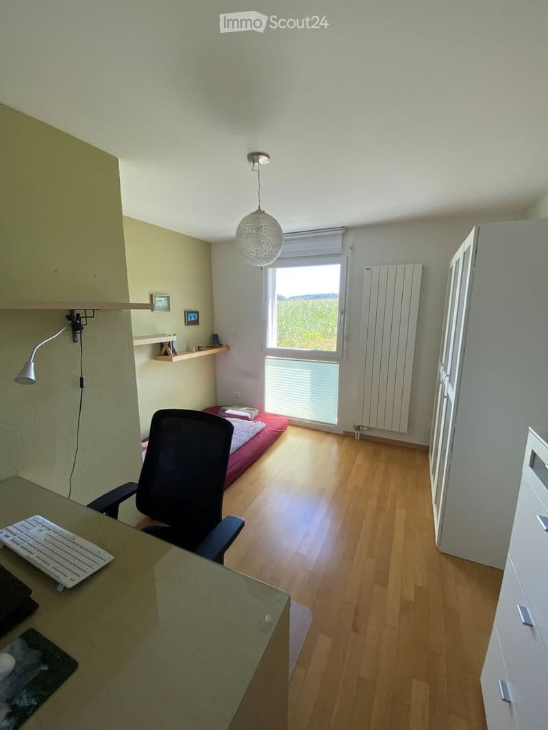 A small, well-lit room with wooden flooring, a bed, a desk with a chair and keyboard, a hanging lamp, and a window with blinds.