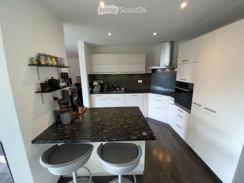 Modern kitchen with white cabinets, granite island, and stainless steel appliances. Two stools are placed at the island, and various kitchen items are visible on shelves and the counter.