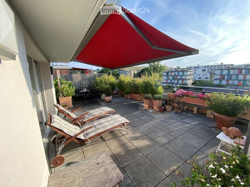 Balcony with lounge chairs, a small table, and several potted plants, featuring a red retractable awning and a view of the city.