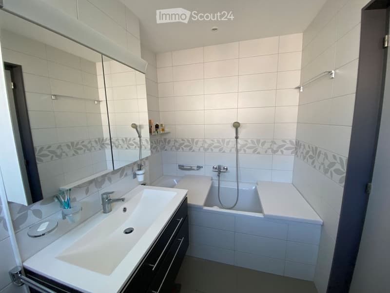 Modern bathroom with white tiles, a large mirror, a sink with a faucet, a bathtub with a showerhead, and a toothbrush holder on the counter.