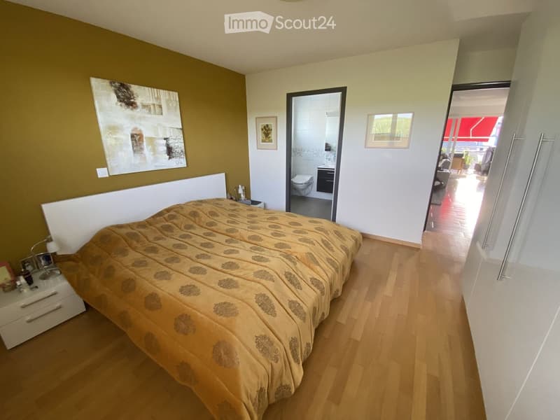 Modern bedroom with wooden flooring, a large bed with patterned bedding, a nightstand with a lamp, and a picture frame on the wall. An open doorway leads to a bathroom with a toilet and sink.