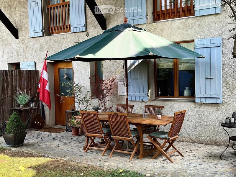 Outdoor patio area with a wooden table, six chairs, and a large umbrella, set against a stone house with shutters and a flag.