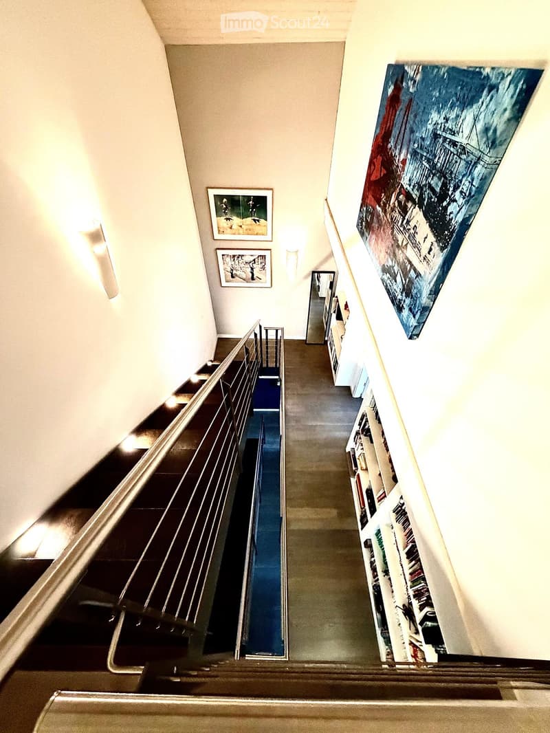 Spacious hallway with a staircase featuring a metal railing, carpeted steps, and a blue runner. The walls are adorned with framed artwork and a large abstract painting. A built-in bookshelf is visible on the right side.