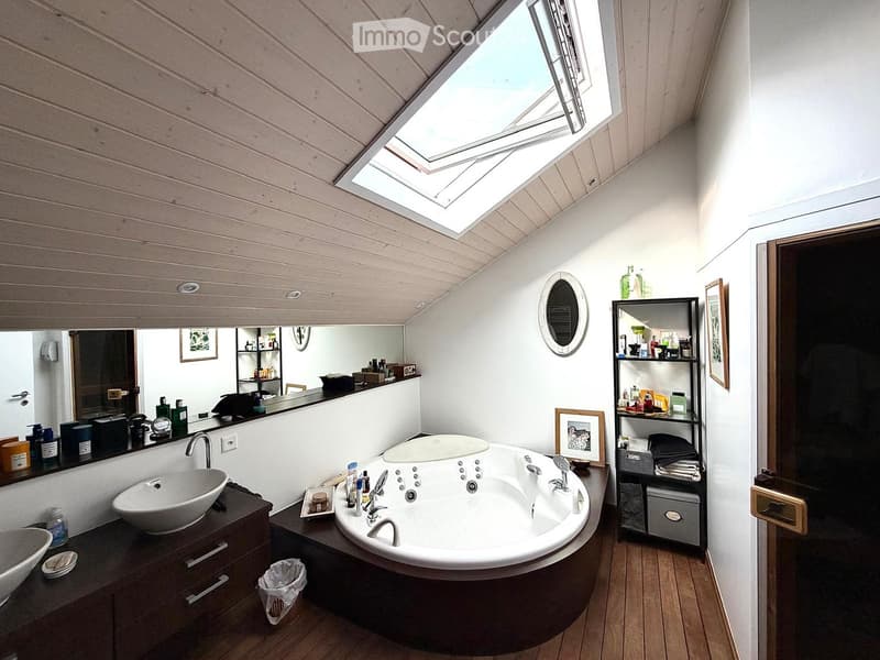 Modern bathroom with a large, round bathtub, a sink, a mirror, shelves, and framed pictures on the walls. The ceiling has skylights, and the floor is wooden.