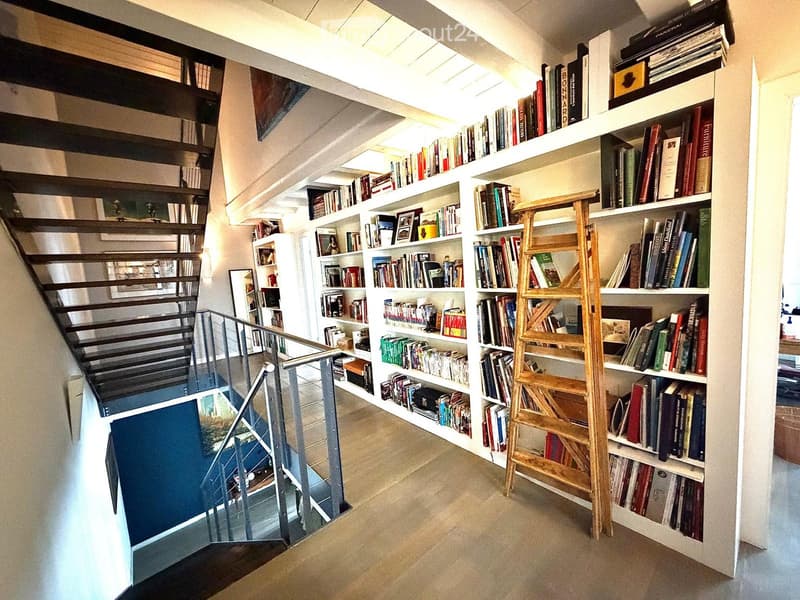 A modern hallway with a staircase featuring a glass railing, white built-in bookshelves filled with books, and a wooden ladder shelf. The space has a clean, minimalist design with neutral tones and soft lighting.