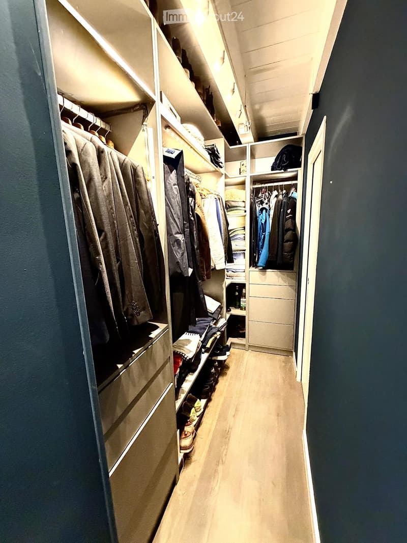 Well-lit hallway with built-in closet units on one side, containing hanging clothes, shelves, and storage compartments. The floor is light-colored wood, and the walls are painted dark blue.