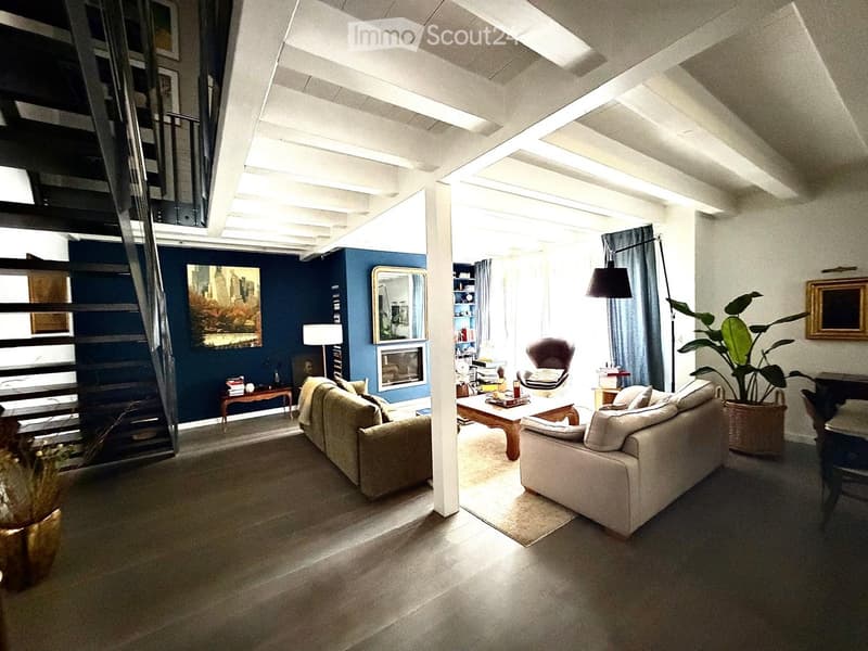 Modern living room with exposed wooden beams, large windows, and a mix of seating including a sofa, armchair, and coffee table. Features a fireplace, bookshelf, and decorative plants.