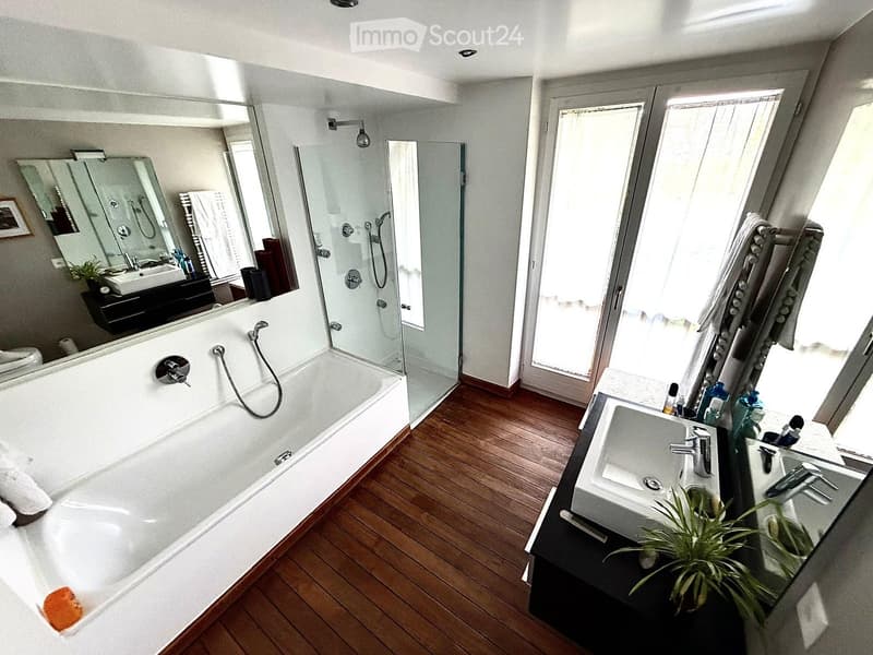 Modern bathroom with a large bathtub, separate shower, double sink, and large mirror. Wooden flooring and natural light from a large window.