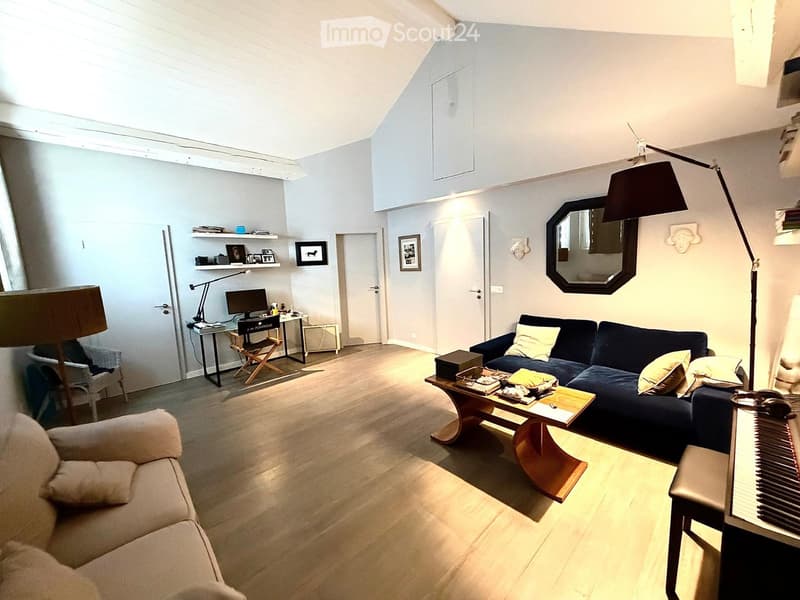Bright living room with wooden flooring, a dark blue sofa, a light-colored armchair, a coffee table, a piano, a desk with a computer, and a mirror on the wall.