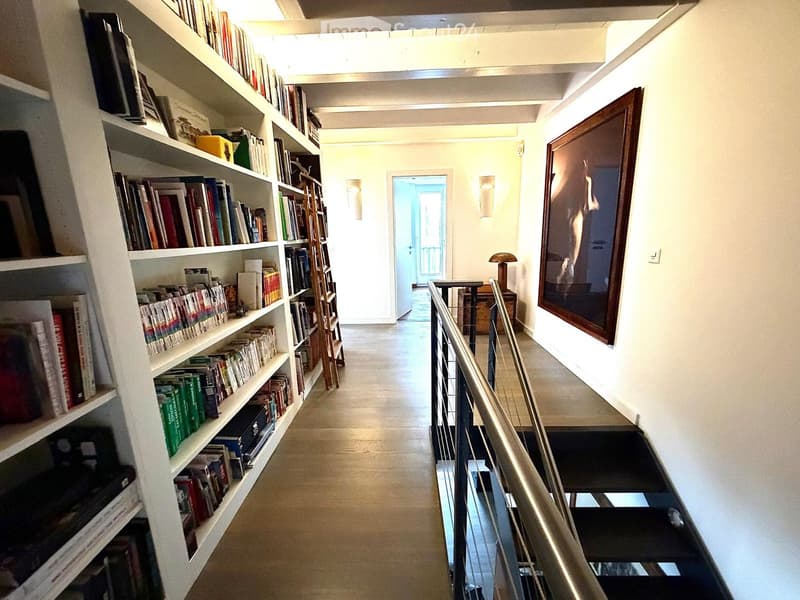 A well-lit hallway with a staircase leading upwards, featuring a bookshelf filled with books on the left and a framed artwork on the right wall.