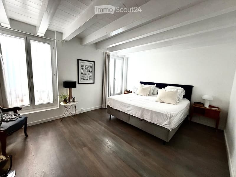 Modern bedroom with a large bed, nightstands, a wooden chair, a small table with a lamp and plant, and large windows with curtains.