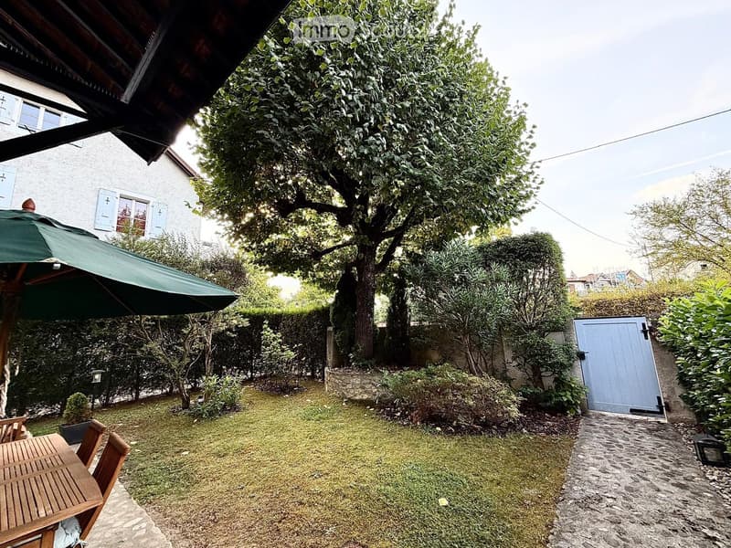 Garden with a wooden table, chairs, umbrella, and a large tree, surrounded by greenery and a stone pathway leading to a gate.