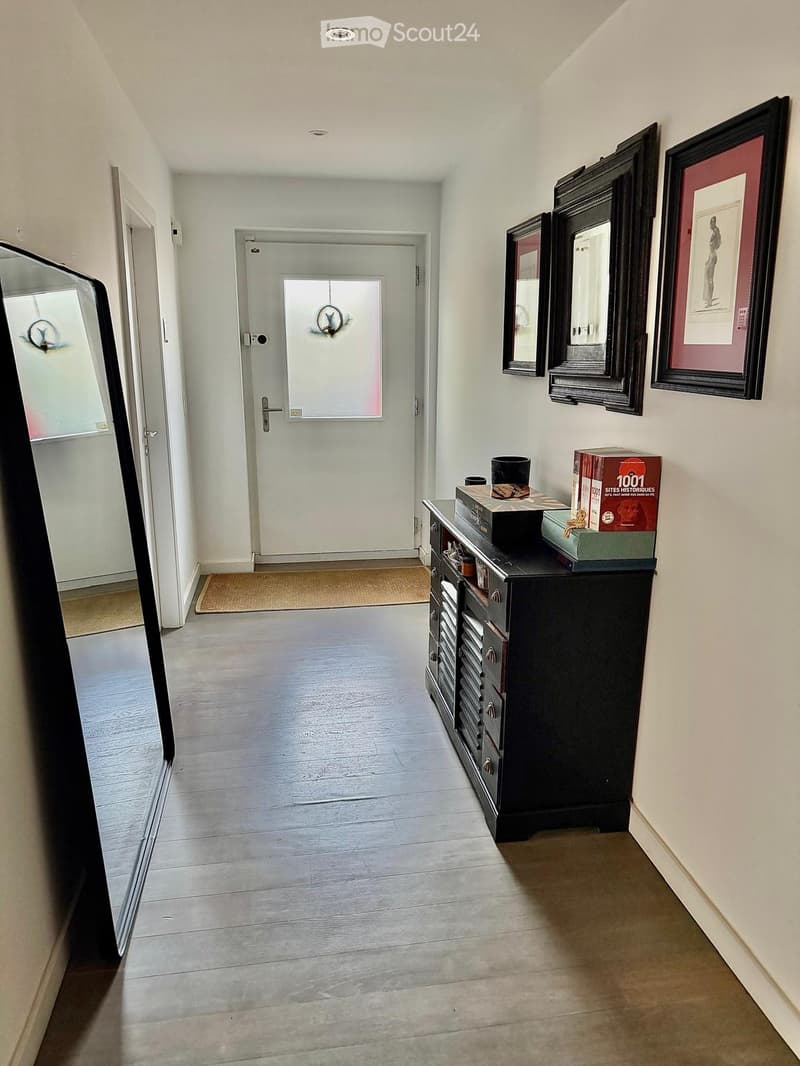 Hallway with wooden flooring, white walls, a black cabinet with drawers, and several framed mirrors and artwork on the wall. A door leads to another room, and a small rug is placed near the entrance.