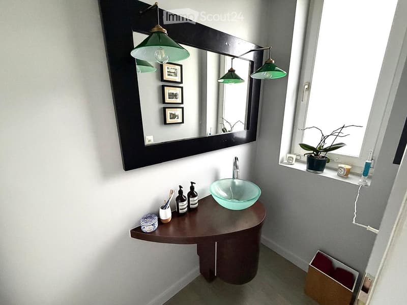 Modern bathroom with a round wooden vanity, glass sink bowl, large mirror with framed pictures, and a window with a small plant on the sill.