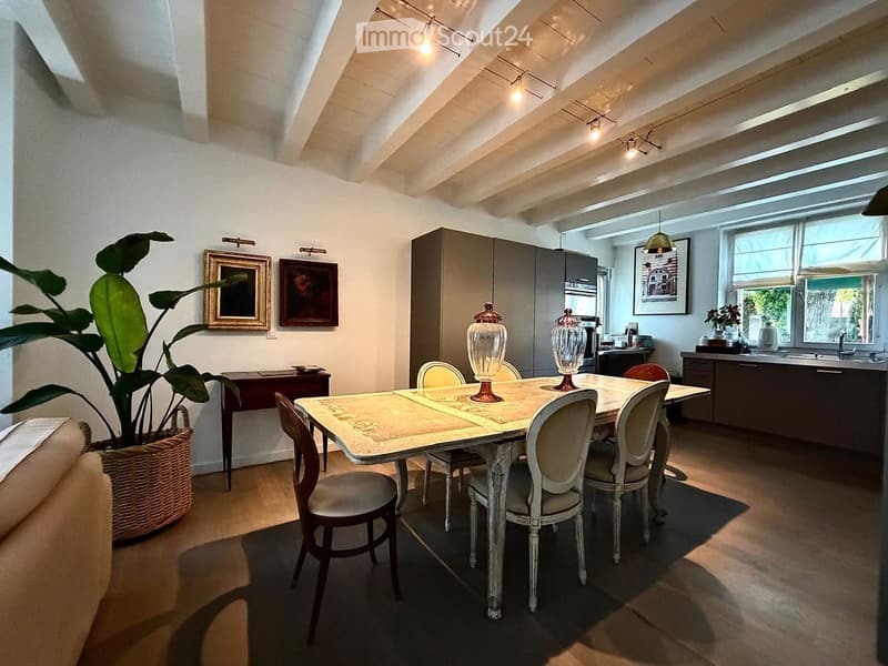Dining area with a table and chairs, a cabinet, and a kitchen area with a sink and faucet. There is a large plant in a basket and framed pictures on the wall.