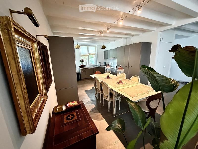A well-lit dining area with a dining table, chairs, and a kitchen area in the background. The room features modern cabinets, a sink, and a large mirror on the wall.