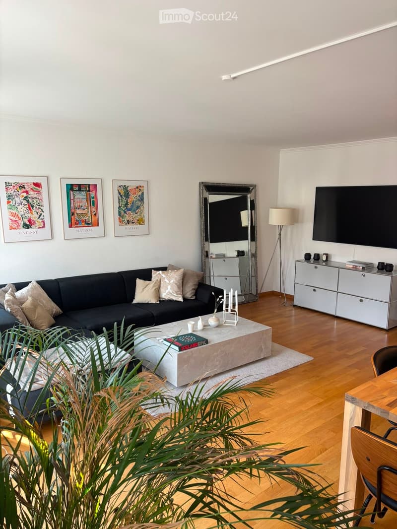 Modern living room with a black sectional sofa, a large coffee table, a TV mounted on the wall, a floor lamp, and a cabinet with drawers.