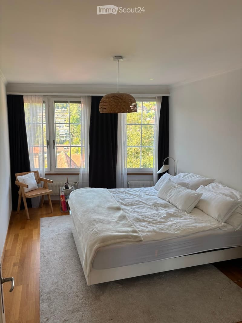 Bright bedroom with a large bed, white bedding, a wooden chair, a small table, and large windows with curtains. The room features a hanging woven lamp and a view of greenery outside.