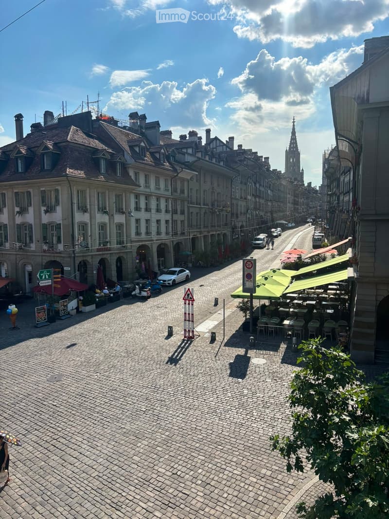 A cobblestone street lined with historic buildings, featuring outdoor seating areas, parked cars, and pedestrians. The street is flanked by multi-story buildings with classic European architecture, including balconies and ornate facades.