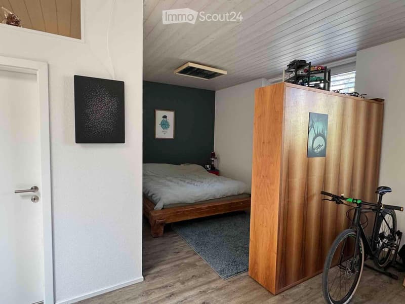Cozy bedroom with a wooden bed, nightstand with lamp, grey rug, bicycle, and wooden cabinet with artwork.