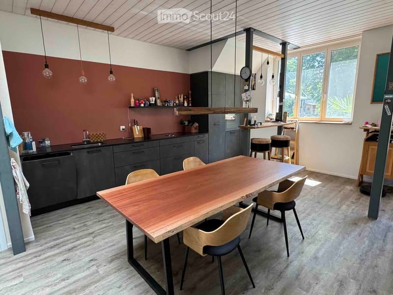 Modern kitchen with dark grey cabinets, a wooden dining table, and hanging pendant lights. The space includes a sink, faucet, and stools near the counter.