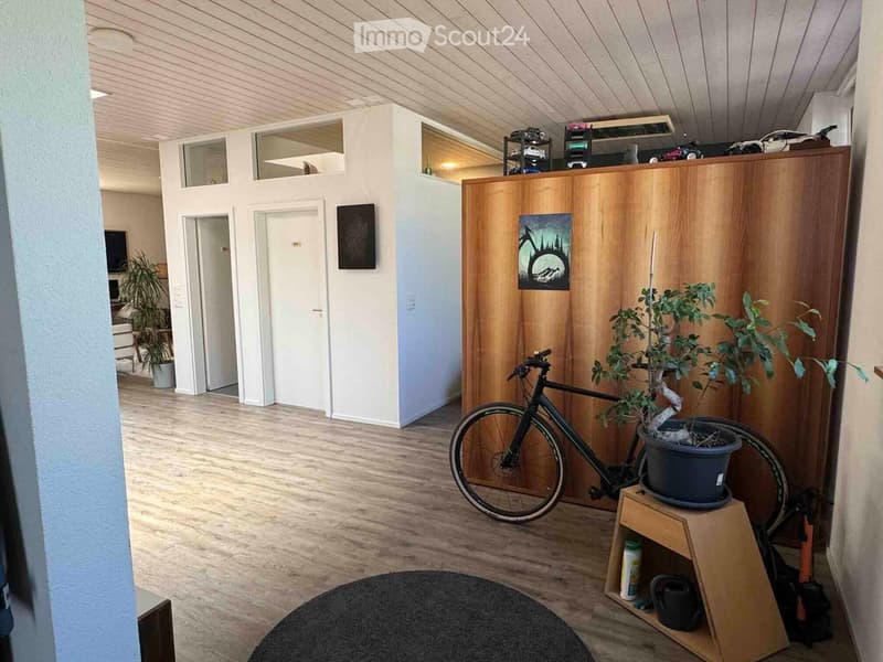 A modern, well-lit room with wooden flooring and white walls. Features include a bicycle leaning against a wooden wall, a potted plant on a small wooden stool, shelves with various items, and a picture frame on the wall.