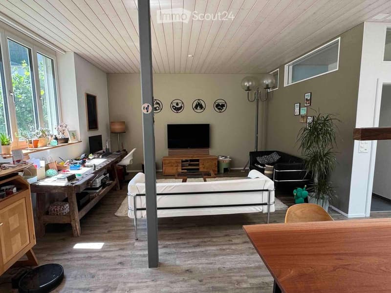 A modern, open-concept living space with a white sofa, black armchair, wooden coffee table, TV, desk, and various decorative items. The room features large windows, wooden flooring, and a slanted ceiling.