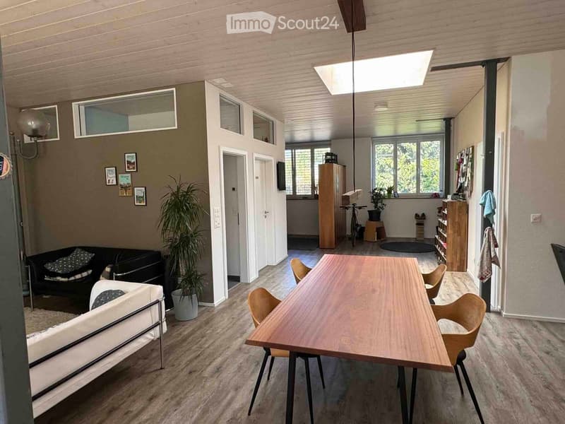 Open-plan living and dining area with wooden flooring, a white sofa, a black couch, a wooden dining table with chairs, and a potted plant. The space features large windows and a modern design.