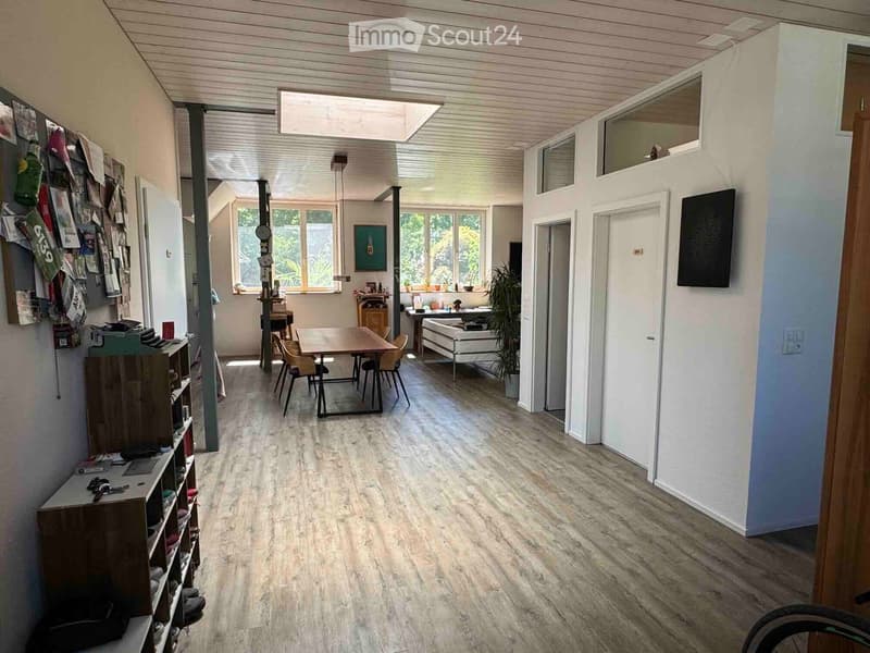Open-plan living and dining area with light wooden flooring, a white sofa, a dining table with chairs, a shelf with shoes and items, and a large screen on the wall.