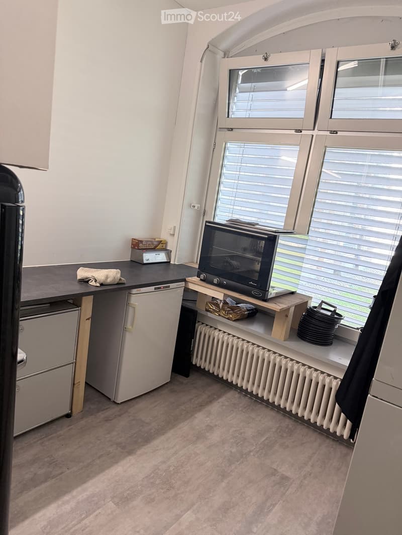 Compact kitchen with a refrigerator, microwave, and small table. The space features large windows with blinds and a radiator beneath them.
