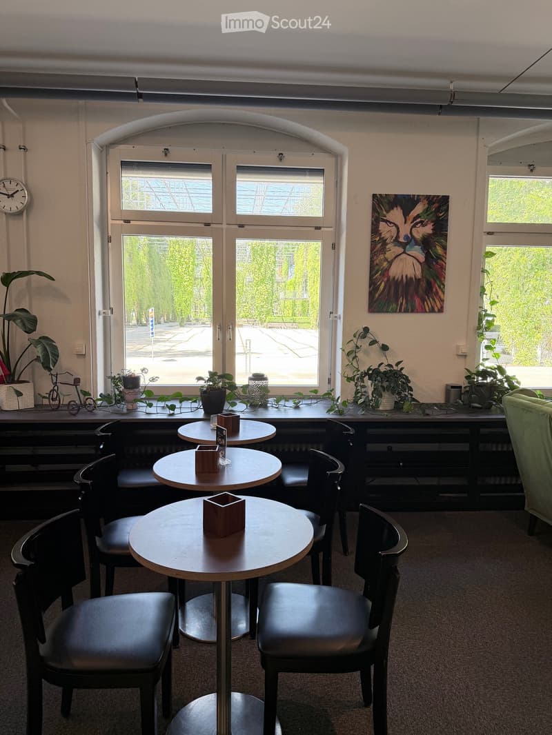Brightly lit room with round tables and chairs, a green sofa, potted plants, and a colorful painting on the wall. Large windows offer a view of greenery outside.