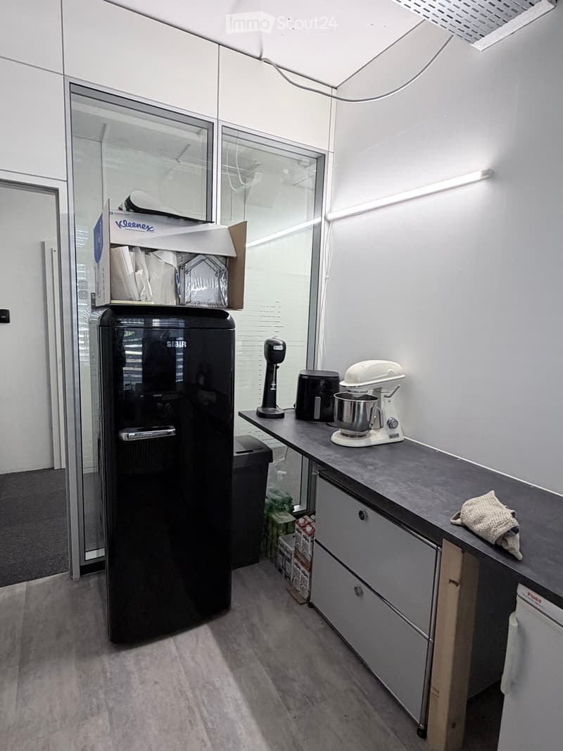Modern kitchen with grey countertops, white cabinets, and stainless steel appliances including a refrigerator, mixer, and toaster. A small box of cleaning supplies is on top of the refrigerator.