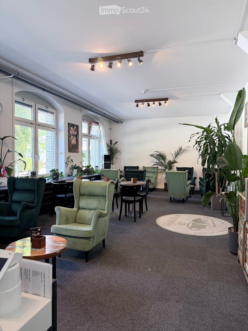 A cozy, well-lit lounge area with green armchairs, small tables, and potted plants, featuring large windows and modern lighting fixtures.