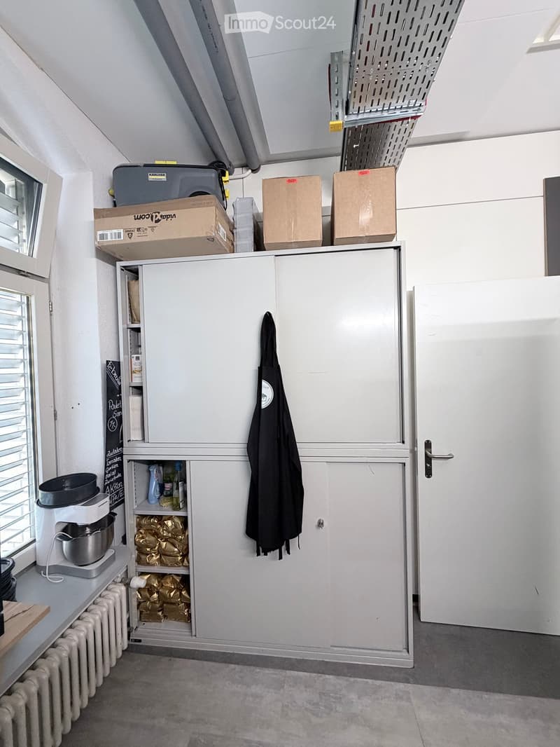 A small, well-organized storage room with white cabinets, a coffee maker on a counter, and various items stored in cardboard boxes and shelves.