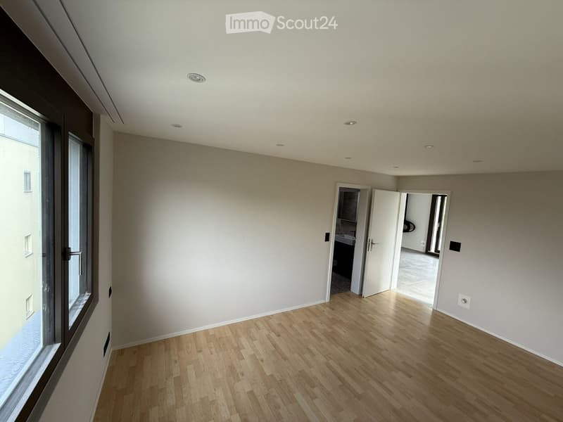 Empty room with light-colored walls, wooden flooring, and recessed ceiling lights. Features a large window and an open doorway leading to another room.
