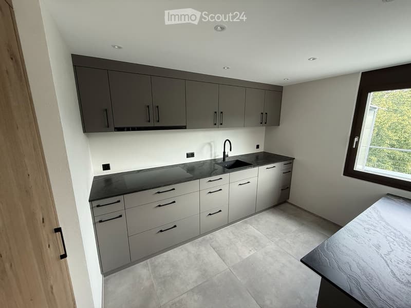Modern kitchen with grey cabinets, white drawers, black countertops, and a large window offering a view of greenery.