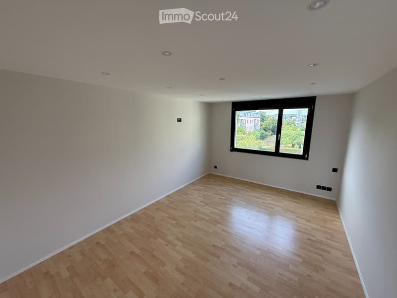 Empty room with light-colored wooden flooring, white walls, and a large window offering a view of greenery and buildings outside.