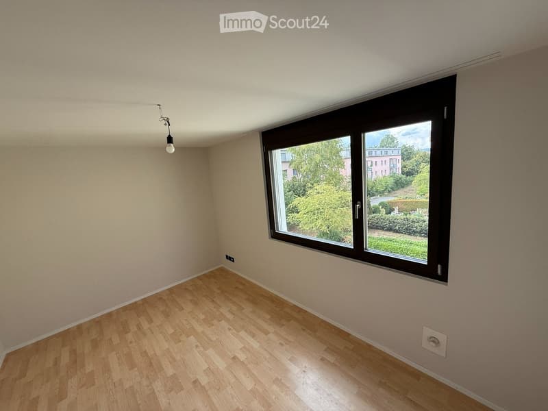 Empty room with light wooden flooring, beige walls, and a large window offering a view of greenery and buildings outside.