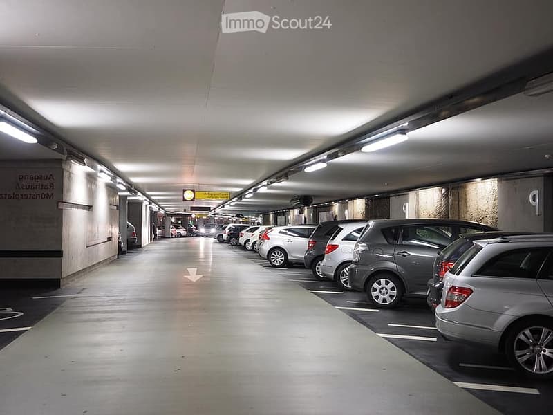 Underground parking area with multiple parked cars, fluorescent lighting, and directional signage.