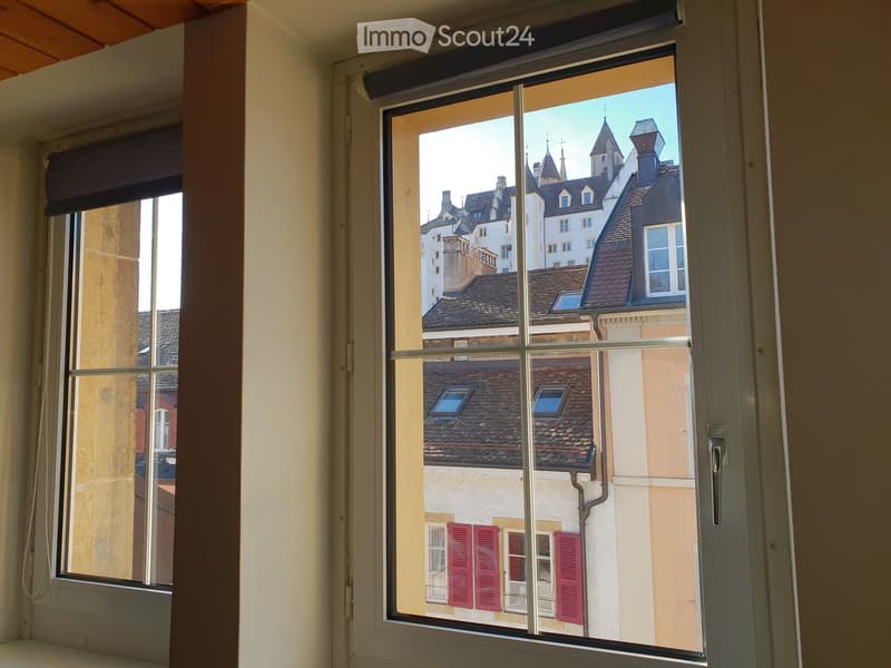 Empty room with large window showing a view of historic buildings with red shutters and tiled roofs.
