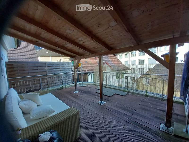 Wooden decked balcony with a wicker sofa, white cushions, and a metal railing, overlooking a residential area with houses and a wooden fence.