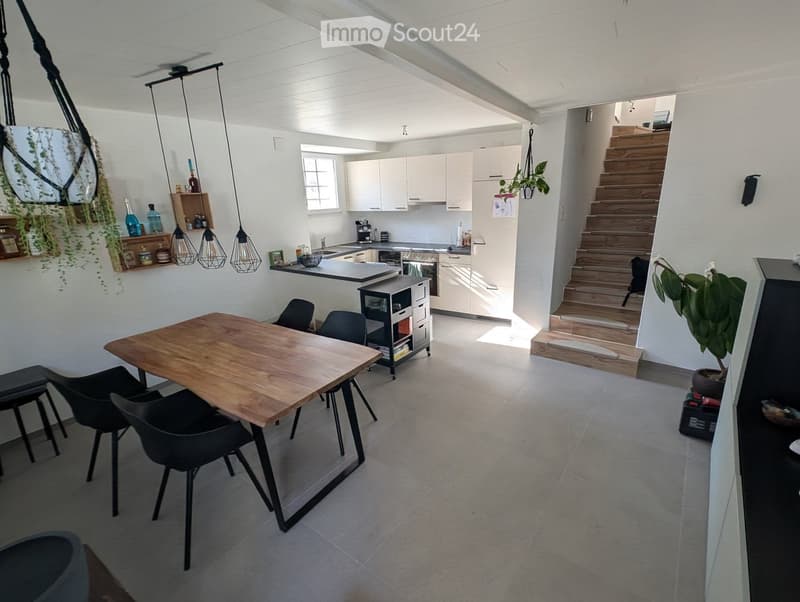 Open-plan living and dining area with a wooden dining table, black chairs, modern kitchen with white cabinets, stainless steel appliances, and pendant lights.