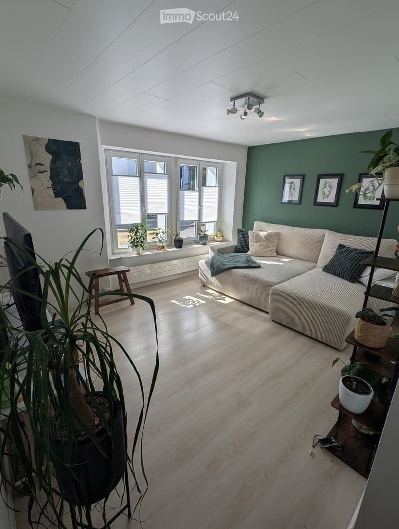 Modern living room with light wooden flooring, large windows, a cream-colored sectional sofa, a small wooden stool, and several potted plants. The room features a green accent wall with framed artwork and a shelf with additional plants.