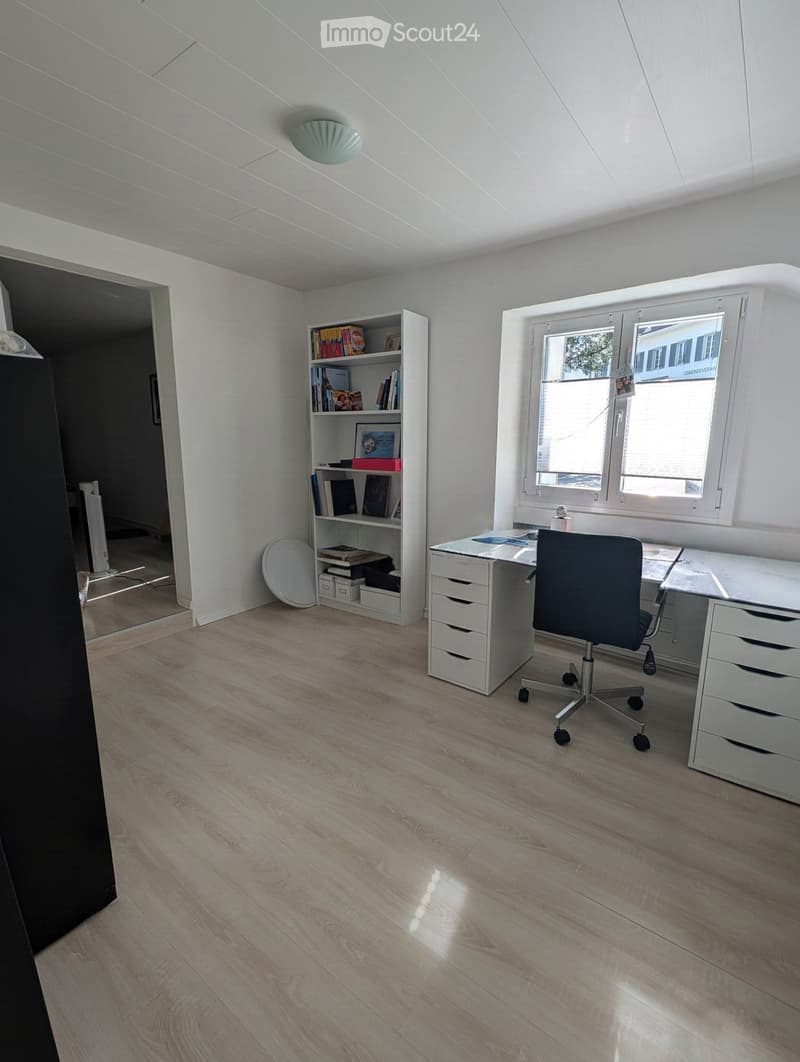 Bright, modern office space with light wooden flooring, white walls, and large windows. Features a white desk with drawers, a black office chair, and a white bookshelf filled with books and decorative items.