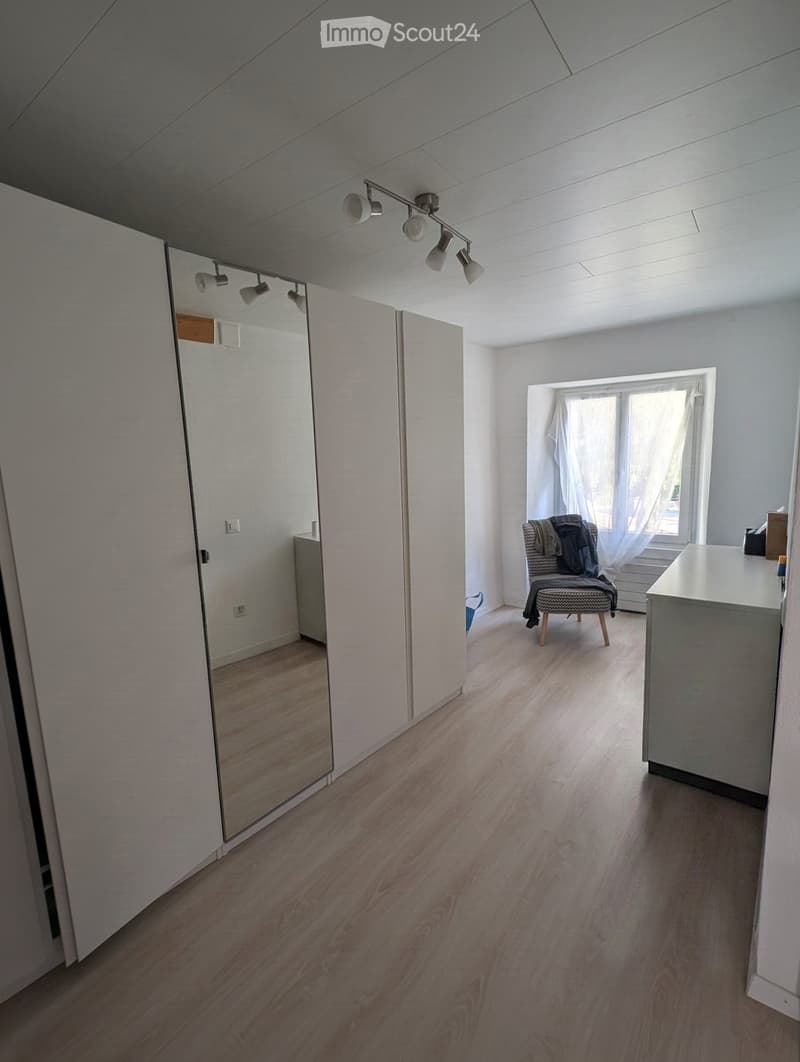 Bright, minimalistic room with wooden flooring, large mirror, and a single chair near a window with sheer curtains.