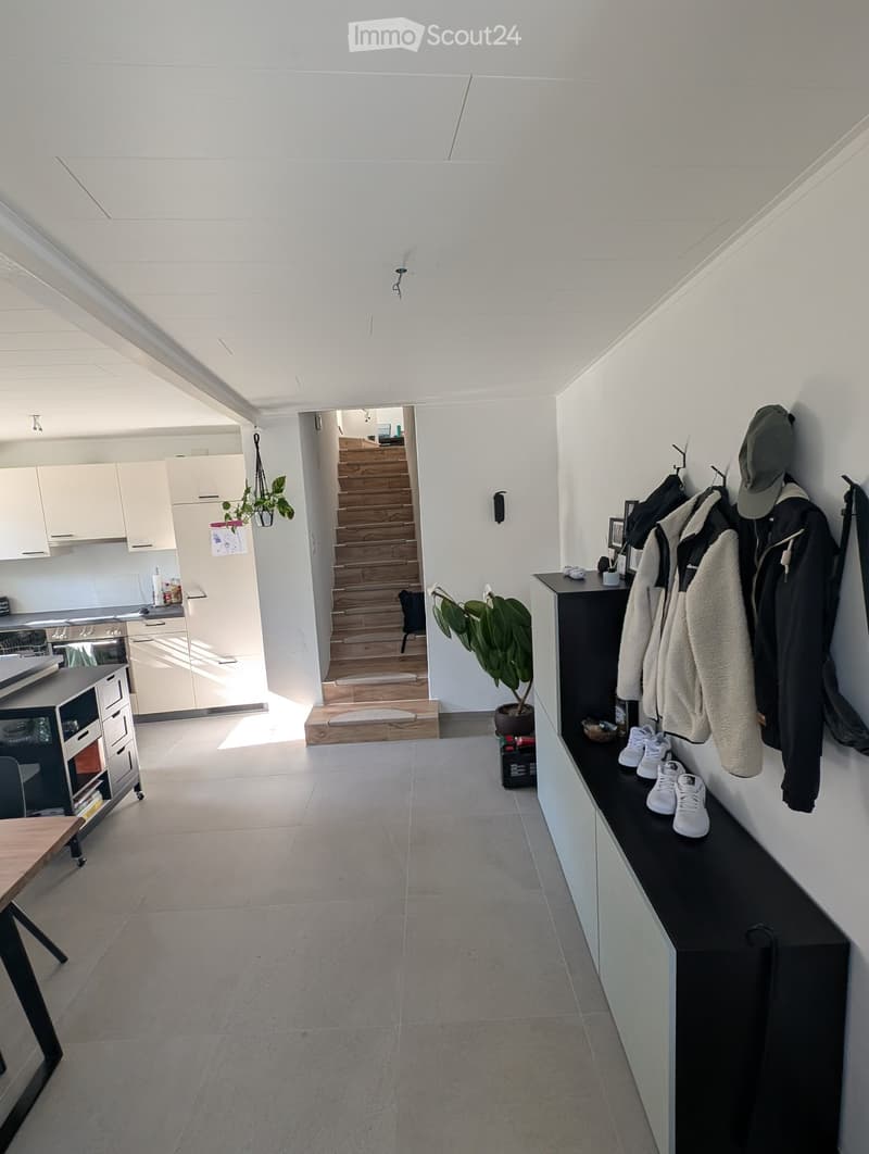 Bright, modern hallway with white walls and ceiling, leading to a kitchen area and staircase. Features a coat rack with jackets and shoes, a small table, and potted plants.