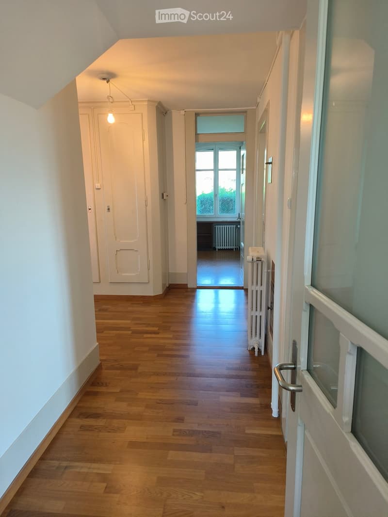 Bright hallway with wooden flooring, white walls, and several doors leading to different rooms. Natural light streams in from a window at the end of the corridor.